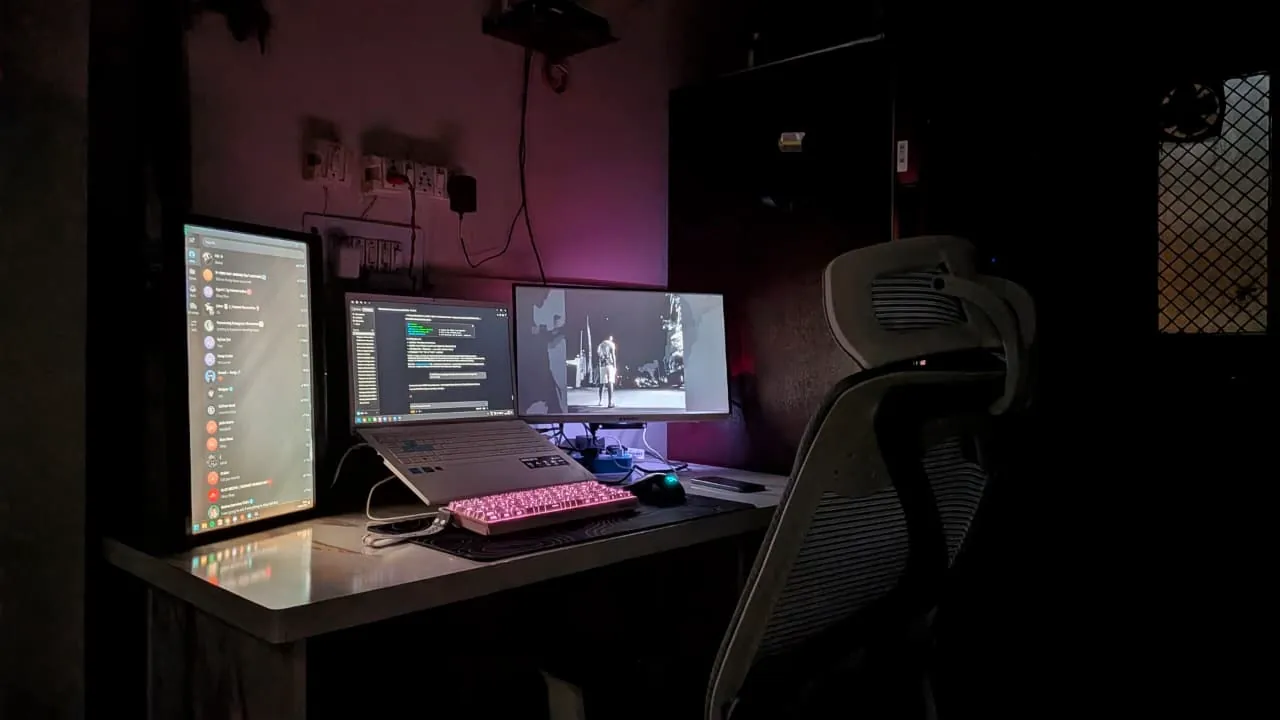 A dark home office at night with three screens and magenta ambient light on the wall